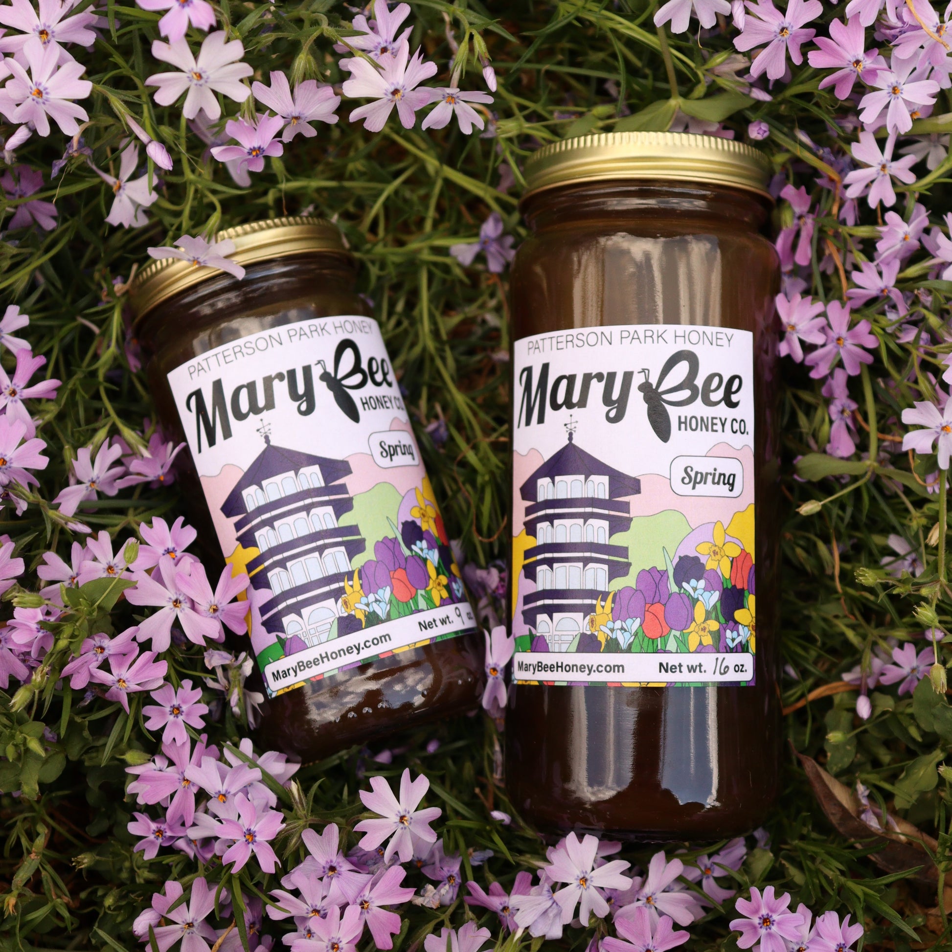 Two jars of dark honey with colorful labels sit in a bed of purple flowers